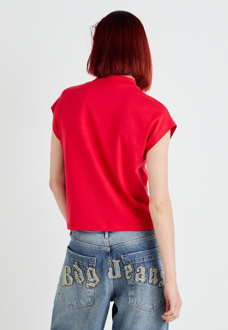 Czerwony krótki top na krótki rękaw z wysoką szyją, w połączeniu z niebieskimi dżinsami z haftowanym napisem "Big Jeans" na tylnych kieszeniach.