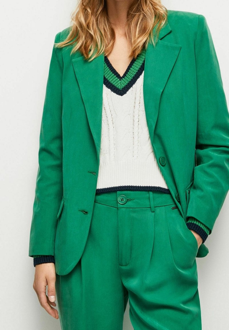 Femme portant un costume vert sur mesure avec un pantalon assorti et un pull en maille blanc bordé de vert et de bleu marine au col et aux poignets.