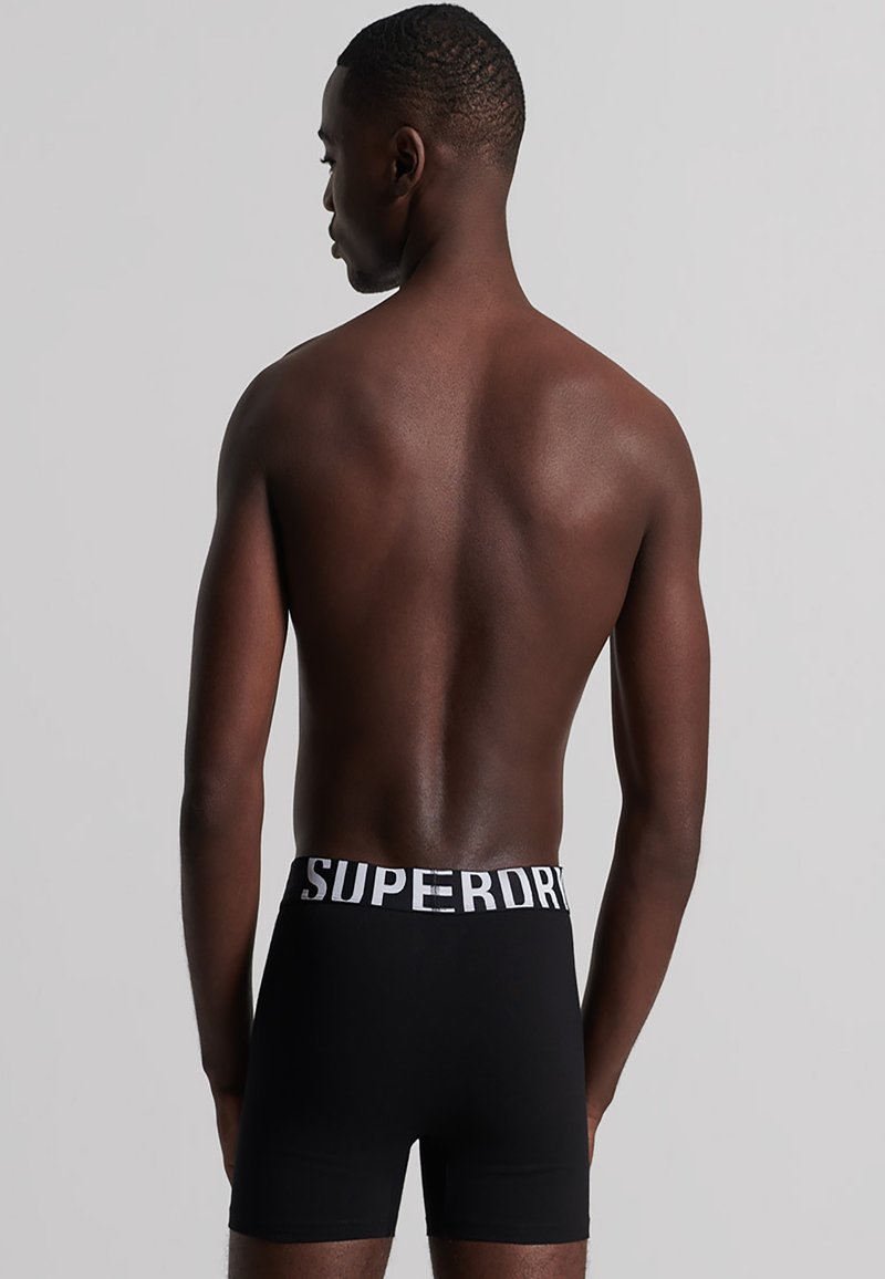 Boxer shorts negros con una cinturilla elástica blanca que presenta las letras "SUPERDRY". El material parece suave y ajustado, sin patrones visibles.