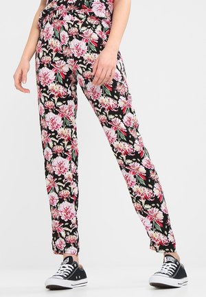 Persoon draagt zwarte bloemenbroek met roze en witte bloemen en zwarte lage sneakers op een witte achtergrond.