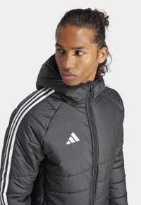 Svart dunjakke med hette, quiltet tekstur, hvit Adidas-logo, og tre hvite vertikale striper på hver skulder.