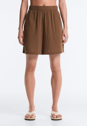 Personne portant un short marron ample texturé et des sandales à plateforme blanches, debout devant un fond uni.