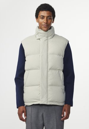 Lichtgrijze puffer vest met een hoge kraag, voorzien van horizontale doorgestikte secties en een rits aan de voorkant. Draagt over een donkergroen lange mouwen shirt.