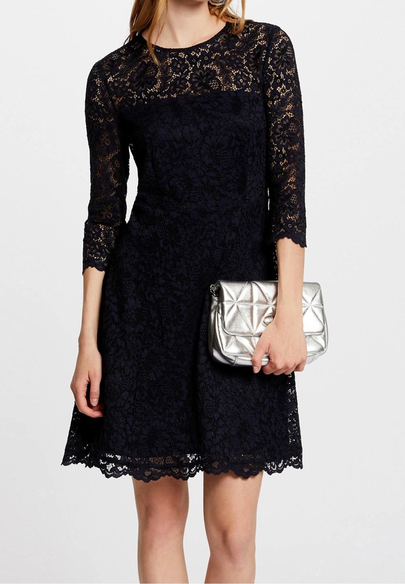 Femme en robe noire en dentelle tenant une pochette argentée matelassée sur un fond blanc uni.