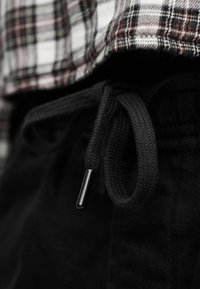 Κοντινή λήψη μαύρου κορδονιού δεμένου σε φιόγκο σε σκούρο παντελόνι με ένα πουκάμισο καρό σε λευκό, καφέ και μαύρο, φορεμένο μέσα στο παντελόνι.