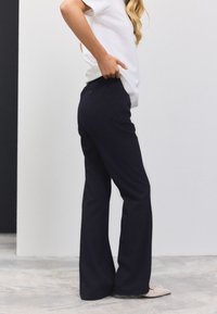 Pantalon évasé bleu marine en tissu lisse, avec une taille dotée d'un bouton à l'arrière, associé à un haut blanc à manches courtes.