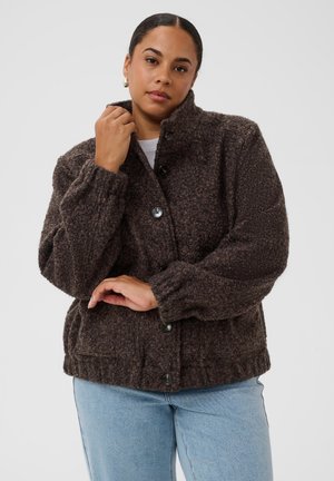 Femme portant une veste marron foncé texturée avec boutons, poignets élastiques et jean bleu clair sur un fond uni.
