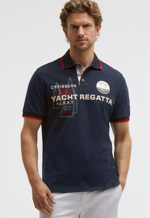 Mann trägt ein marineblaues Polohemd mit rotem Rand, gesticktem Schriftzug "Yacht Regatta", Segelgrafik und weißen Shorts vor hellem Hintergrund.
