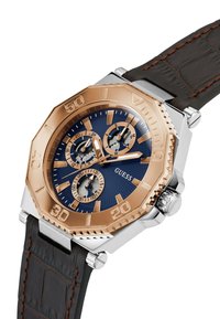 Reloj Guess con un bisel octagonal de oro rosa, esfera azul marino, tres subesferas y una correa de cuero texturizado marrón con costuras marrones.