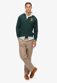 Mørkegrøn langærmet polo med gul logo, hvid skjorte med krave under, beige bukser og brune sko. Glat tekstur, afslappet pasform.