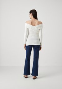 Balta, off-the-shoulder palaidinė su prigludusia forma, ilgomis sijonomis, su ruching detalėmis nugaroje, derinama su tamsiai mėlynomis platiomis džinsais ir sandalais.