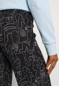 Svarta byxor med vita abstrakta linjeteckningar som visar mönster av ansikten och former. Ribbat tyg och detalj med elastisk midja.