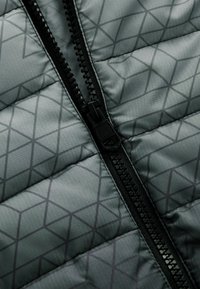 Veste matelassée grise avec un motif géométrique. Présente une fermeture éclair noire et un tissu texturé, mettant en valeur une finition lisse et brillante.