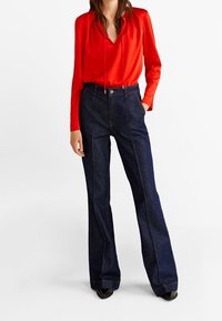 Blusa roja de satén de manga larga con escote en V combinada con jeans de mezclilla oscuros de talle alto y pierna acampanada, con costuras laterales en contraste.
