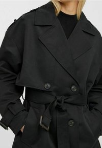 Czarny płaszcz trench z podwójnym zapięciem, szerokimi klapami i paskiem w talii. Wykonany z ciężkiego materiału o gładkiej teksturze.
