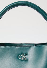 Geantă din piele teal cu o textură netedă, având un logo argintiu "CK" și o mână robustă, evidențiind cusături precise pe margini.