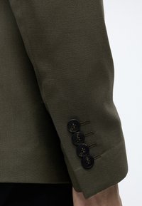 Gros plan sur la manche d'une veste de costume vert foncé avec quatre boutons noirs le long du poignet, portée sur le bras d'une personne.
