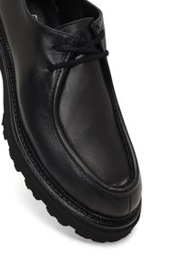 Zapato de piel negra con puntera redondeada, costuras elevadas y suela de goma negra texturizada. Cuenta con cordones en la parte superior para el ajuste.