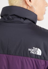 Purple and black nylon jacket with high collar, featuring a white branded logo on the back, showcasing smooth texture and modern design.