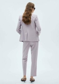 Costum personalizat de culoare lavandă deschisă, cu un sacou structurat și pantaloni slim fit, având o deschidere la spate și sandale negre elegante.