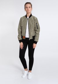 Olivgrüne Bomberjacke mit Reißverschluss, schwarzen Rippbündchen, seitlichen Taschen und einem Patch an der unteren Vorderseite. Getragen über einem weißen Hemd und schwarzen Hosen.