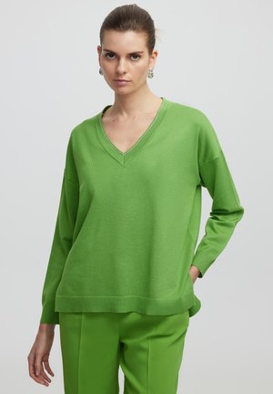 Suéter de punto verde brillante con cuello en V y mangas largas. Presenta un dobladillo y puños acanalados. Combinado con unos pantalones verdes a juego.