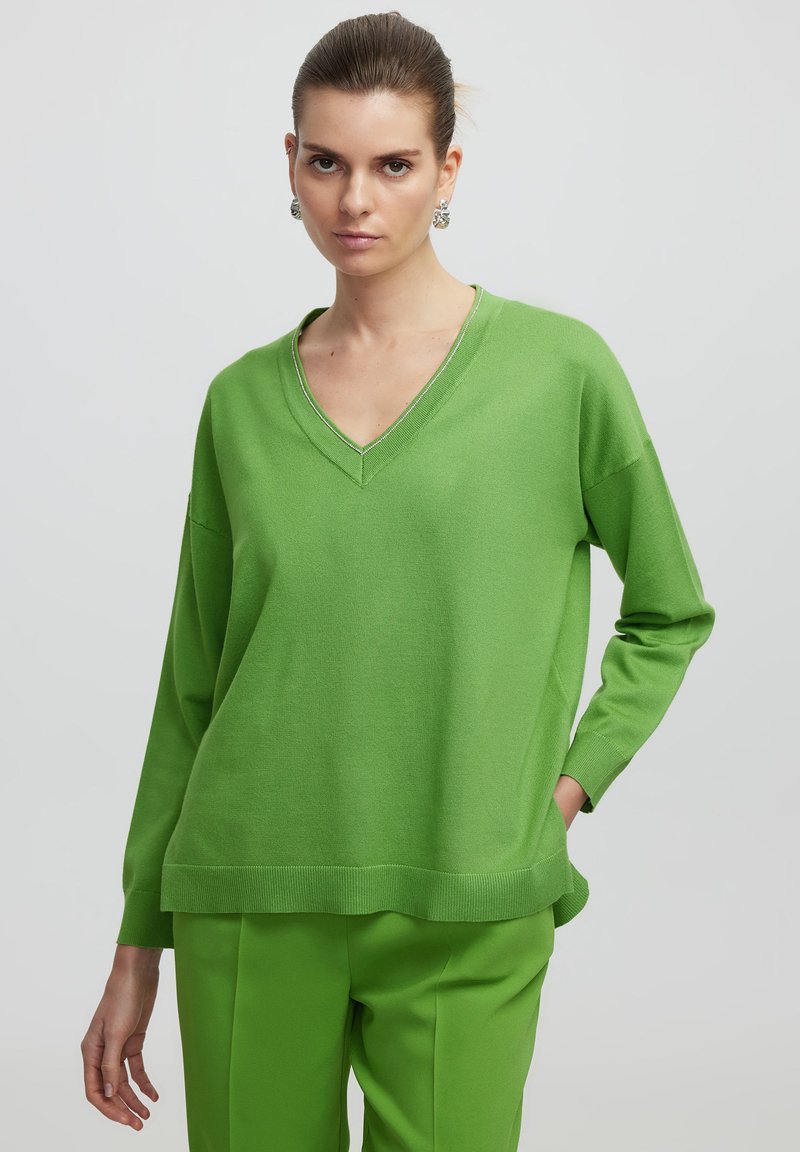 V-Ausschnitt, langärmliger Pullover aus leuchtend grünem Strick. Mit einem gerippten Saum und Bündchen. Getragen mit passenden grünen Hosen.