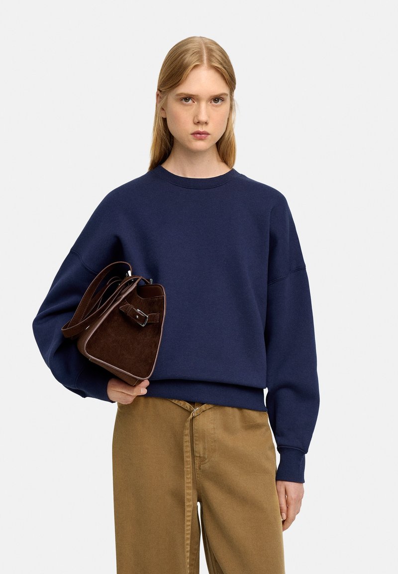 Granatowy sweter z opadającymi ramionami, połączony z beżowymi spodniami typu wide-leg. Brązowa torebka z zamszu o wyraźnym kształcie i z klamrą.