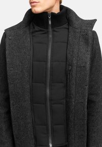 Schwarze, gestepptes Innenjacke mit hohem Kragen, unter einem strukturierten grauen Wollmantel getragen. Verfügt über Knöpfe und mit Reißverschlüssen versehenen Taschen.