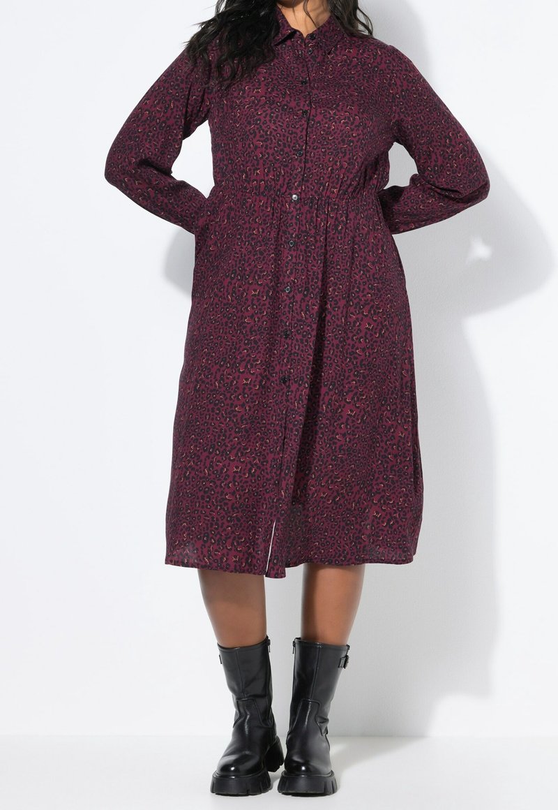 Femme portant une robe violette à manches longues, boutonnée, imprimé léopard avec des bottines noires épaisses mi-mollet, debout les mains derrière le dos.