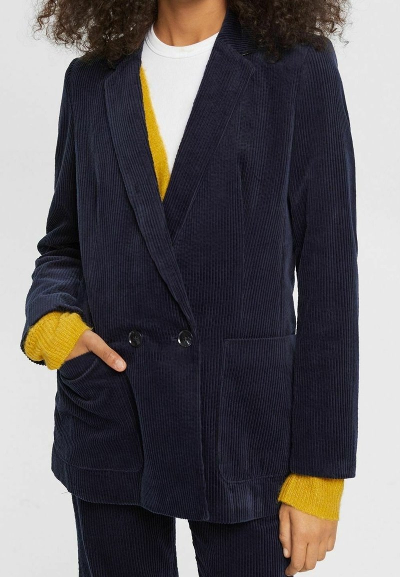 Femme portant un blazer croisé en velours côtelé bleu marine avec poches plaquées sur un haut blanc et des manches de pull jaune apparentes.