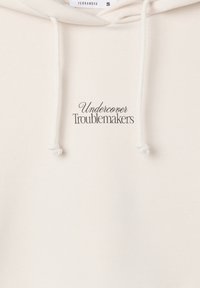 Felpa con cappuccio color crema, realizzata in tessuto morbido, con cappuccio regolabile tramite coulisse e la scritta "Undercover Troublemakers" ricamata in nero.