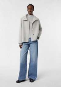 Giacca grigia strutturata con colletto e due bottoni frontali. Abbinata a jeans di denim leggeri e larghi di colore blu. Calzature nere completano l'outfit.