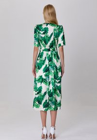 Robe à manches courtes, imprimé de feuilles tropicales vertes, avec une taille à nouer, longueur midi et texture de tissu lisse. Chaussures à talons blancs visibles.