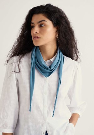 Svijetloplavi šal od mekanog materijala, prebačen oko vrata, uparen s bijelom teksturiranom košuljom na kopčanje i ležerno stiliziranom kosom.