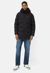Mand med en sort hættetrøje, blå jeans og hvide sneakers, stående med en neutral ansigtsudtryk på en hvid baggrund.