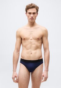 Νεαρός άνδρας με σγουρά μαλλιά, φοράει μπριφ μπλε ναυτικό με μαύρη μέση, στέκεται μπροστά σε απλό ανοιχτό γκρι φόντο.