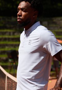Lys grå polo-shirt med ribbet krave og en struktureret overflade, der har et lille sort logo på venstre bryst, båret på en tennisbane.