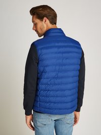 Blå puffervest med horisontale quiltede seksjoner, høy krage og en glatt tekstur. Båret over en mørk langermet skjorte.