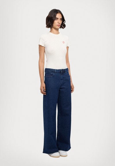 Hvid kortærmet top med bølgede kanter og et lille lyserødt logo, parret med vide mørkeblå jeans og hvide sneakers.