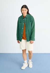 Grønn denimjakke med to brystlommer, brukt over en oransje skjorte og hvite stripete shorts; paret med kremfargede sneakers.