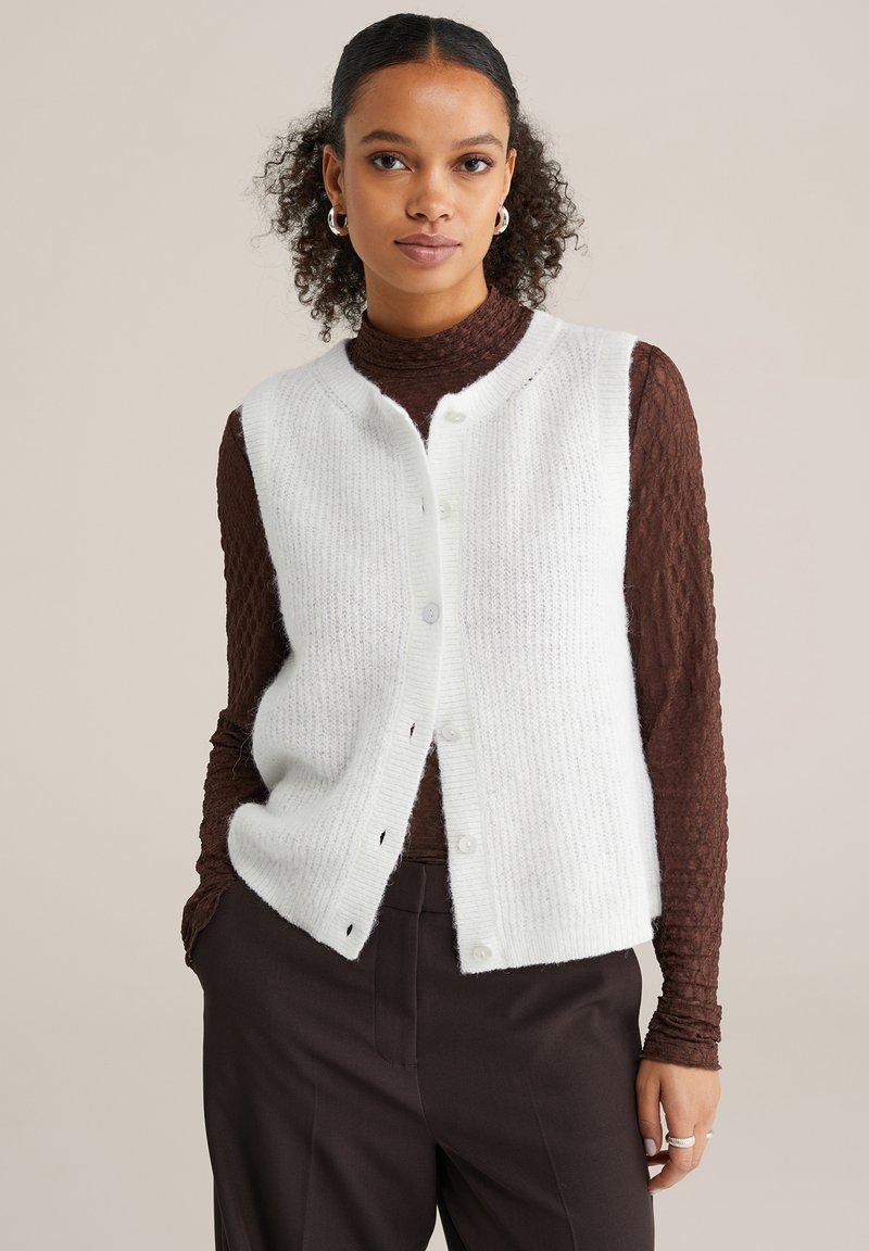 Žena v bielej zapínacej pletenej veste cez hnedý štruktúrovaný dlhý rukáv a tmavohnedé nohavice, stojaca s rukami v vreckách.