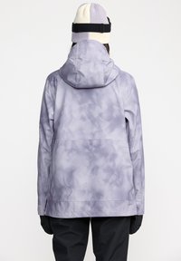 Chaqueta con un patrón de tie-dye púrpura y capucha, con una textura suave y brillante, y bolsillos laterales con cremallera, llevada con un gorro de punto bicolor.
