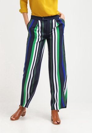 Vrouw die broek met wijde pijpen draagt met verticale strepen in groen, blauw, zwart en wit, een gele top en bruine open-toe hakken.