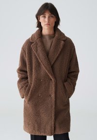 Brauner, doppelt geknöpfter, flauschiger Mantel mit für den Schnitt typischen Revers, seitlichen Taschen und strukturiertem Stoff. Der Mantel reicht bis unter die Hüfte.