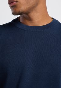 Sinih ribbed neule, jossa on pyöreä kaula-aukko, teksturoitu kangas ja rento malli. Kultaketju näkyvissä kaulassa.