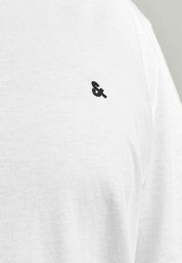 Vit bomullströja med en liten svart broderad ampersand-symbol på bröstet. Slät textur med rundad halsringning.