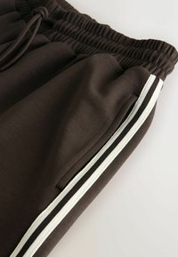 Brązowe dresy z elastycznym pasem, z bocznymi kieszeniami oraz kontrastowymi białymi i czarnymi paskami wzdłuż zewnętrznych szwów.