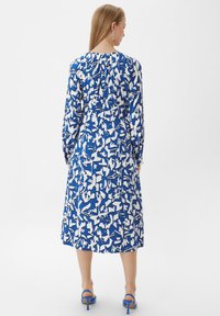 Blau-weiß gemustertes Kleid mit langen Ärmeln, Knöchellänge und gerafftem Ausschnitt. Getragen mit blauen Absatzsandalen.