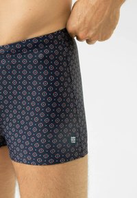 Marineblauwe zwembroek met een cirkelpatroon met rode en witte accenten. Materiaal lijkt glad en rekbaar. Merkenlabel zichtbaar.